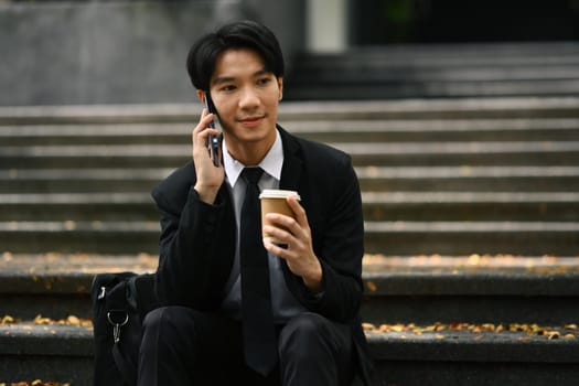 Image of asian businessman in formal wears talking on mobile phone while sitting on the stairs outdoors in urban area.