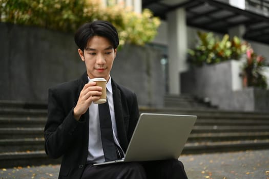 Young businessman in formalwear sitting outside of office in urban city and using laptop.