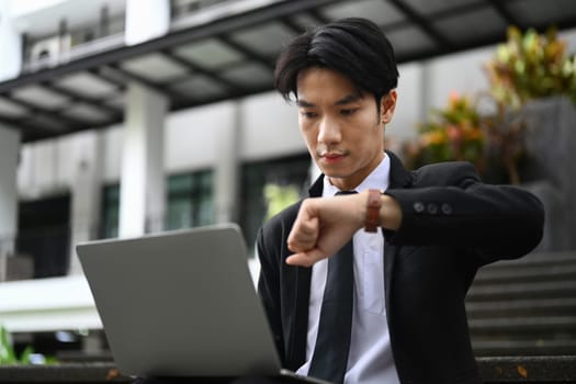 Young businessman using laptop and checking time on wristwatch for his next appointment.