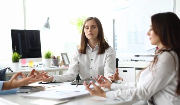 Portrait of gorgeous businesswoman sitting at modern workplace and closing tender eyes in calmness and satisfaction. Beautiful woman doing special meditation pose. Business teamwork concept