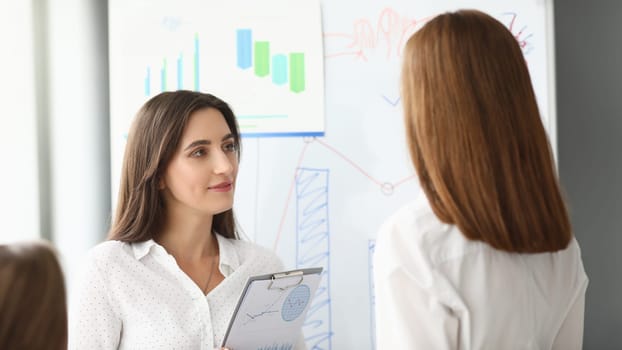 Portrait of joyful businesswoman standing at modern workplace and holding important paper tablet with charts and graphs and looking at somebody with calmness. Accounting office concept