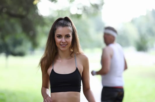 Portrait of beautiful fit female posing on camera outdoors. Morning park run for strong body. Man watching pretty girl from back. Sport and active lifestyle concept