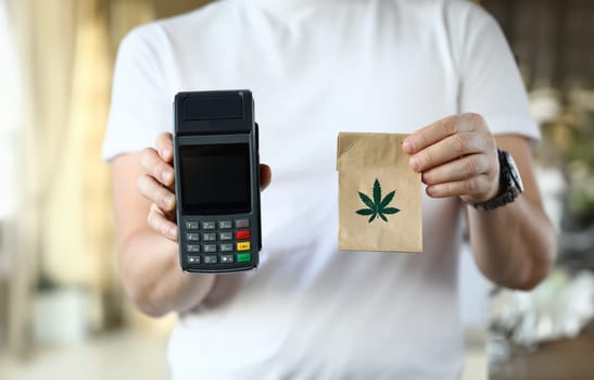 Close-up of courier holding paper package with traditional meds. Cannabis in bag with leaf symbol. Cashier machine for fast payment. Nfc technology counter and remedy concept