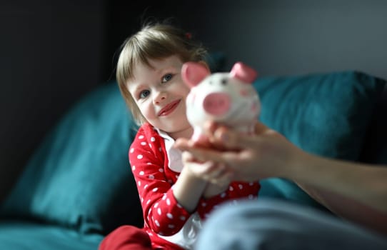 Portrait of lovely little kid holding pink piggy bank. Smiling light haired girl wearing red dress in dots. Charming toddler posing on camera. Money and saving up for toy concept