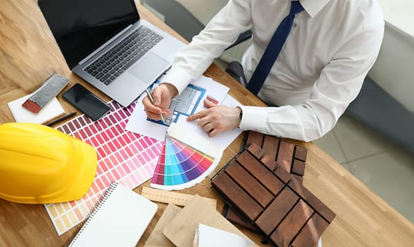 Top view of person sitting in office and working with house plan. Renovation in apartment. Palette with colour samples. Yellow helmet on wooden table. Architect and interior design concept