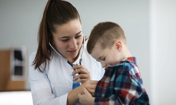 Portrait of cheerful little boy on examination by pediatrician. Doctor listening to breathing or action heart of ill patient with stethoscope. Healthcare and medicine concept