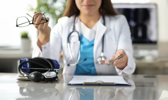 Close-up of therapist hands holding glasses and pen. Tonometer and documents on table. Doctor office interior on background. Healthcare and medicine concept