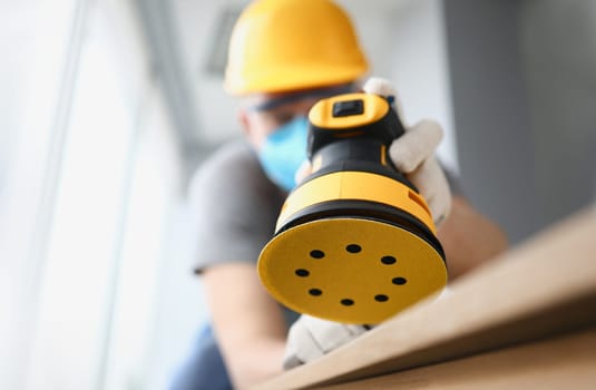Close-up of professional craftsman working with electrical yellow sander. Worker wearing protecting uniform and face mask. Renovation and construction site concept