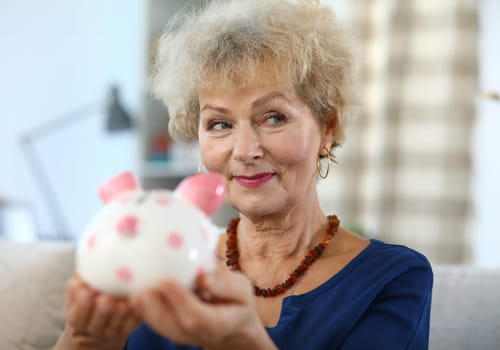 Close-up of attractive elderly woman holding piggy bank for money. Colourful container for coins and cash. Beautiful female indoors. Saving for future and economy concept