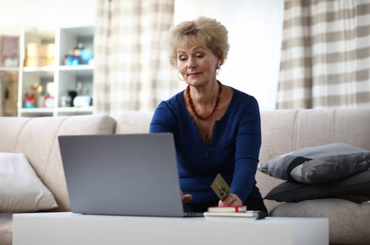Portrait of beautiful elderly woman sitting on sofa at home. Charming female shopping via internet using laptop. Lady holding credit card. Modern technology and spare time concept