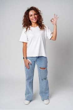 Cheerful young woman in a white shirt and ripped jeans making an okay gesture in studio