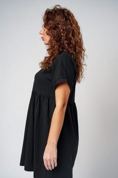 Curly-haired woman in a black dress poses sideways against a neutral backdrop close up