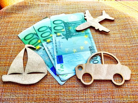 Three wooden shapes representing transport are arranged alongside euro bills, illustrating cost comparisons for travel options.