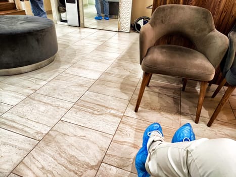 A person is waiting in a doctor's clinic hallway, sitting with feet in blue shoe covers on tiled flooring