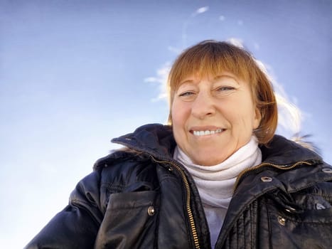 A cheerful middle aged woman in a winter coat taking selfie on nature outdoors in sunny day with blue sky