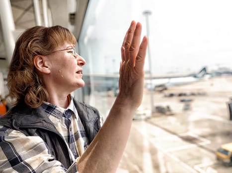 A middle aged woman stands by the airport window, waving goodbye while her flight is about to depart in a reflective moment.