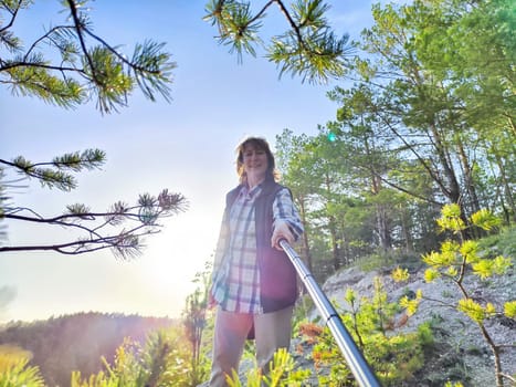 A middle-aged female tourist blogger takes a selfie on a travel. A woman takes a selfie with a selfie stick in forest and blue sky on background
