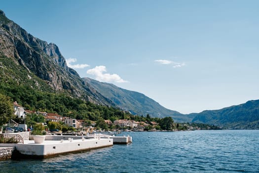 White pier on the shore of the bay against the backdrop of ancient houses at the foot of the mountains. Dobrota, Montenegro. High quality photo