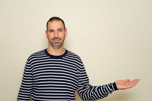 A bearded Hispanic man in his 40s wearing a striped sweater extending his hand to the side in a gesture of offering or outstretching, holding it palm forward. Isolated on beige background
