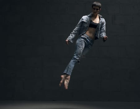 Girl with shot haircut in jeanswear jumping gracefully in dark room full-length shot