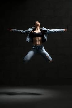 Ballet dancer with shot haircut in unbuttoned jeans shirt jumping gracefully in dark room