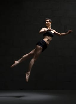 Ballet dancer with shot haircut in top and shorts jumping gracefully in dark room
