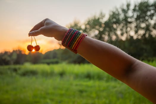 A hand beautifully adorned with colorful bracelets gently holds cherries against a stunning sunset backdrop