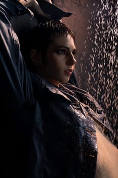 Brunette in wet jeans shirt standing under water in dark room profile portrait