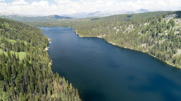 A breathtaking aerial view of a tranquil lake enveloped by lush, dense forest, with distant mountain ranges under a clear, blue sky. A perfect depiction of nature's untouched beauty.