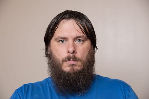 Close-up portrait of a man with a thick beard and wet hair, wearing a blue shirt, against a neutral background.