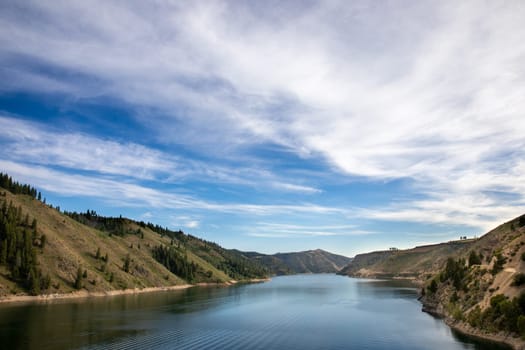 A beautiful river flows between rolling hills under a vast blue sky. The serene landscape evokes peace and tranquility, showcasing the natural beauty and calmness of the environment.