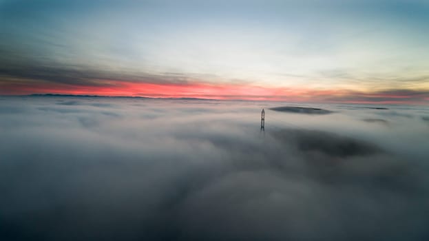 Serene aerial view of misty sunrise with power line emerging through fog over landscape, creating a tranquil and mystical atmosphere.