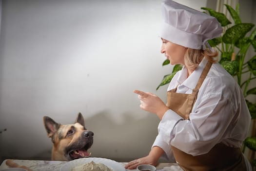 An elderly woman dressed as a cook shares a light-hearted moment with her shepherd dog while preparing flour-based dishes.