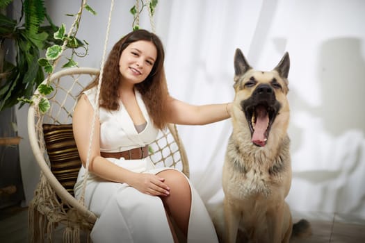 A woman sits on a swing while petting her yawning German Shepherd in a relaxed indoor atmosphere filled with plants.
