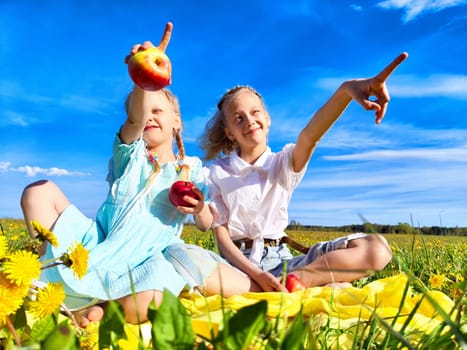 Two girls sit on a blanket in a lush flower field, joyfully holding apples and pointing at the sky during a beautiful sunny day.
