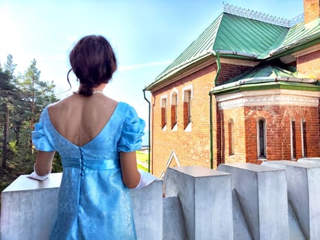 A young lady in a blue gown stands on a balcony, admiring the view from an ancient castle under clear skies.