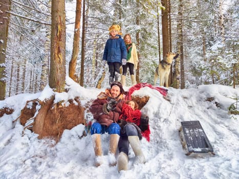Joyful family ethnic dress with shawls and earflap hats and dog in winter forest in Russia carnival Maslenitsa. Tourists in Shrovetide in spring. Mother, father, son, daughter having fun in snow