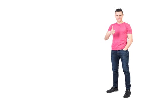 Full body of satisfied young male model with dark hair smiling and showing thumb up while standing against white background with hand in pocket and looking at camera