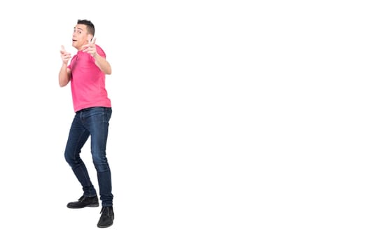 Full body of young funny male model with dark hair in pink t shirt and jeans looking and pointing at camera against white background