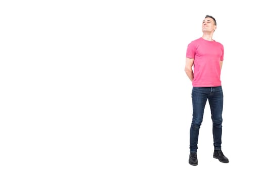 Full length of self assured young man in casual clothes standing in white studio with hands behind back and looking away proudly