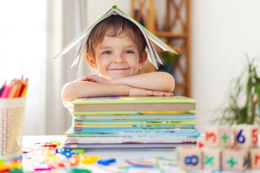 A smiling child with a book opened over their head like a roof, resting on a stack of books. The child is at a desk surrounded by colored pencils, toys, and number blocks.