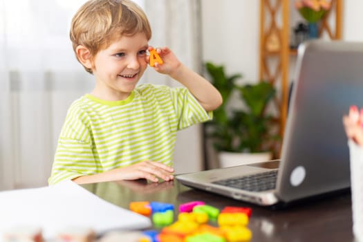 A young boy wearing a green striped shirt looking at a laptop while holding a toy letter. The table is filled with colorful toy letters and paper, creating an educational atmosphere.