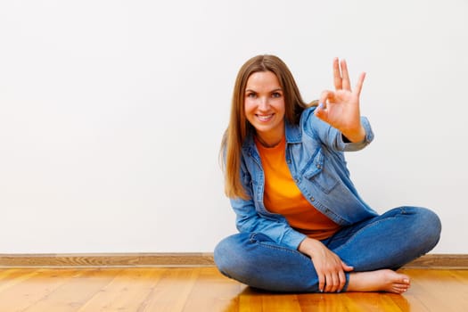 A smiling woman in a casual denim outfit sitting cross-legged on a wooden floor against a white wall, showing an OK hand gesture. Studio shot suitable for greeting card, poster, and banner design.
