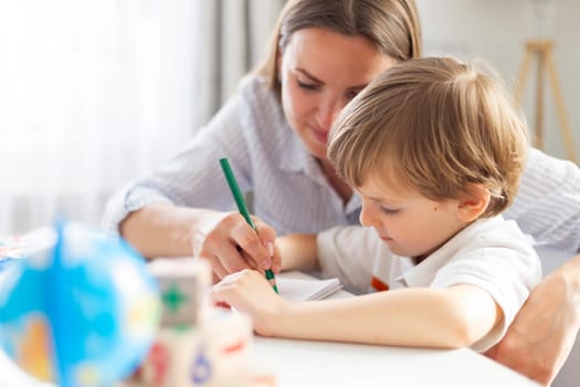 Mother assisting young boy with writing at a table, surrounded by educational toys and materials in a home setting. Indoor scene emphasizing learning and parent-child interaction.