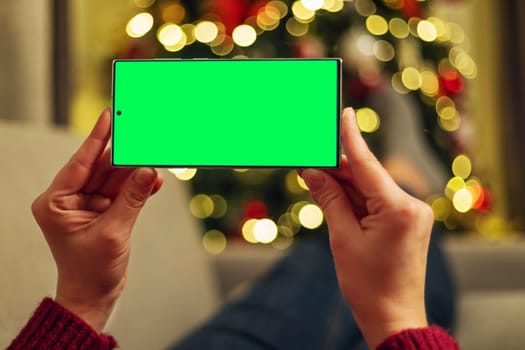 Hands holding a smartphone with a green screen in front of a blurred Christmas tree background. The device is positioned horizontally with bokeh lights in the background.