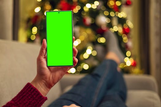 A person holding a smartphone with a green screen while reclining on a couch near a Christmas tree decorated with lights. The background features blurred holiday decorations.