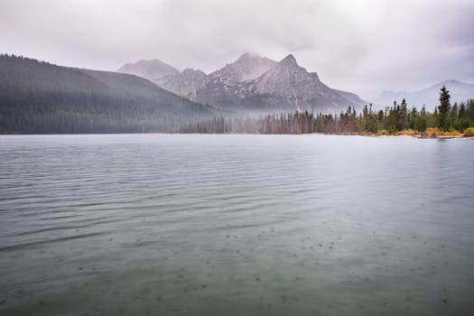 A tranquil mountain lake surrounded by mist-covered forests and towering peaks in a serene, overcast atmosphere, showcasing the untouched beauty of nature.