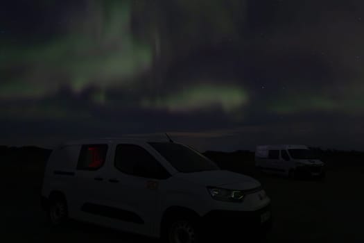 A white van is parked in a field at night. The sky is filled with auroras, creating a serene and peaceful atmosphere