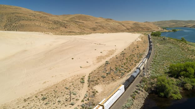 Aerial view of a freight train winding through a vast desert landscape beside a tranquil river. The scene captures the essence of solitude and adventure in a remote, natural environment.