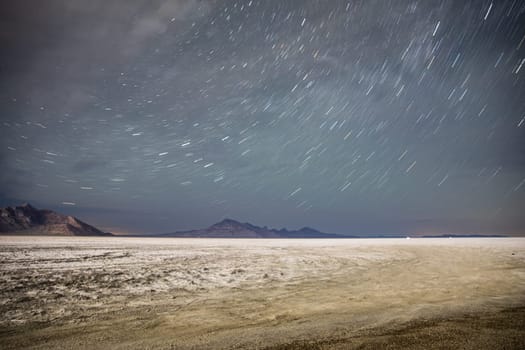 A mesmerizing view of the starry night sky with long exposure over a wide, barren desert. The serene desert terrain beautifully contrasts the dynamic star trails in the sky.